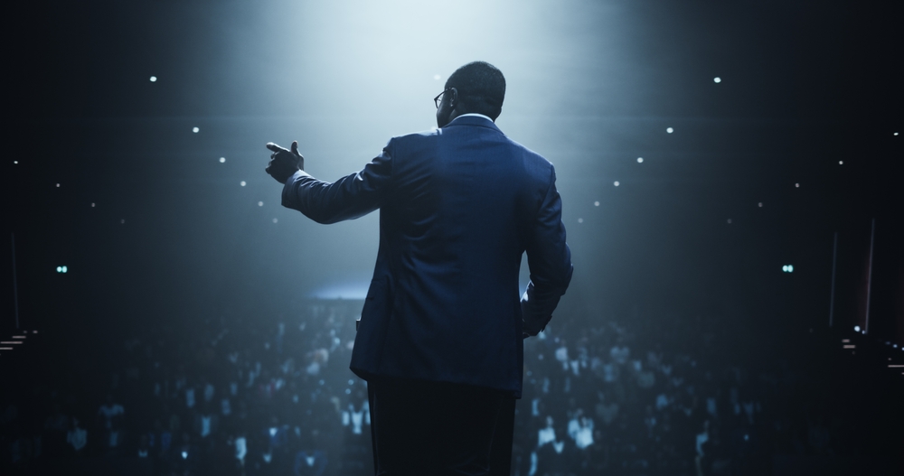 Líder em palco iluminado por holofote discursa para grande plateia em auditório — motivação, liderança e negócios